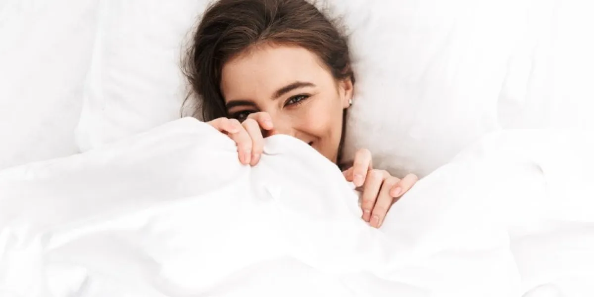 photo from above of cute woman 20s smiling, while lying in bed on white pillow after sleep or nap