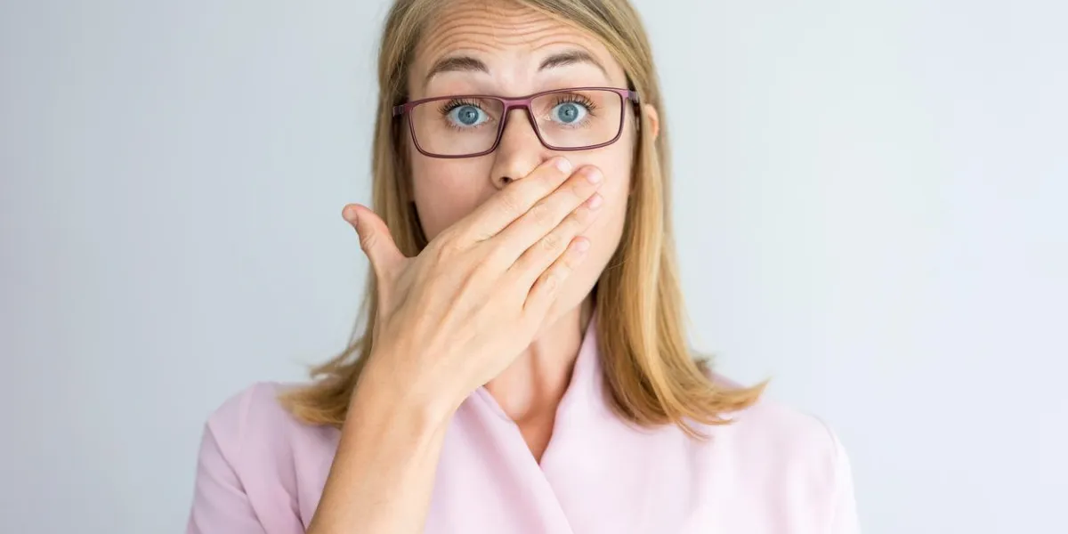 close-up of shocked young caucasian businesswoman wearing pink blouse and glasses covering mouth with hand surprise and confusion concept
