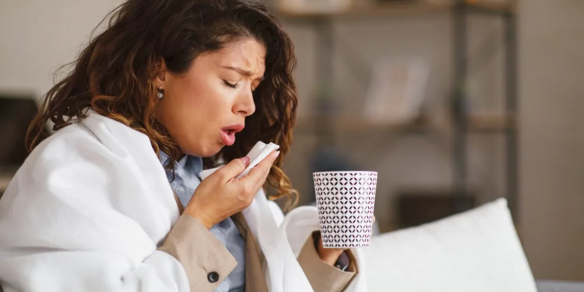 young woman coughing use paper tissue holds a cup of tea and is wrapped in a blanket at home