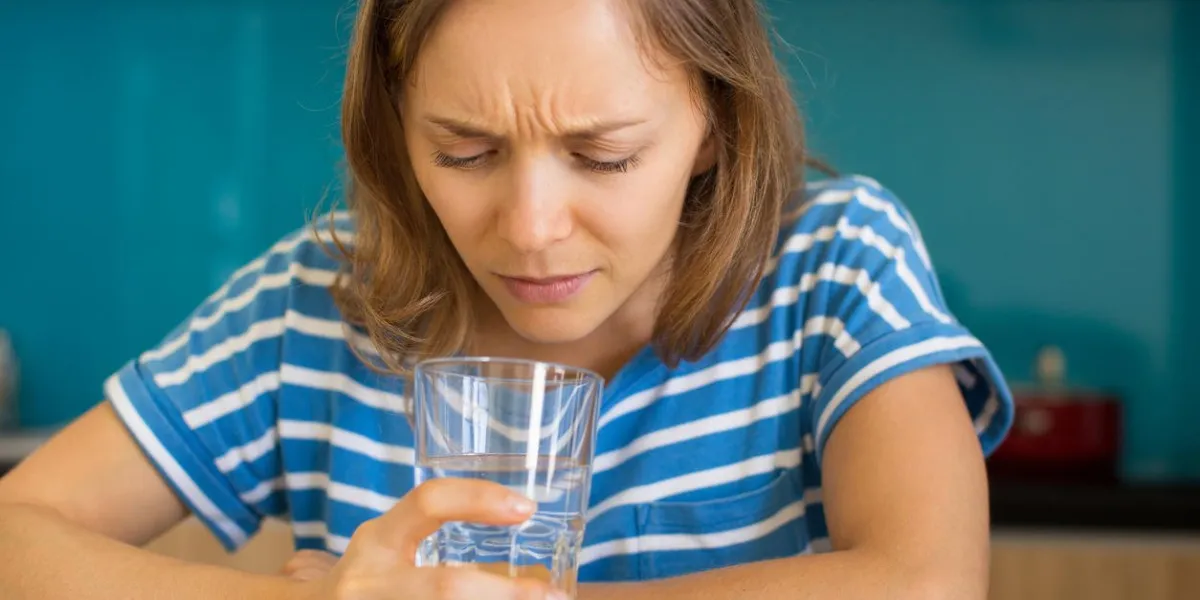 closeup portrait of dissatisfied young beautiful woman looking into glass of water and sitting at table in kitchen bad quality drinking water concept front view