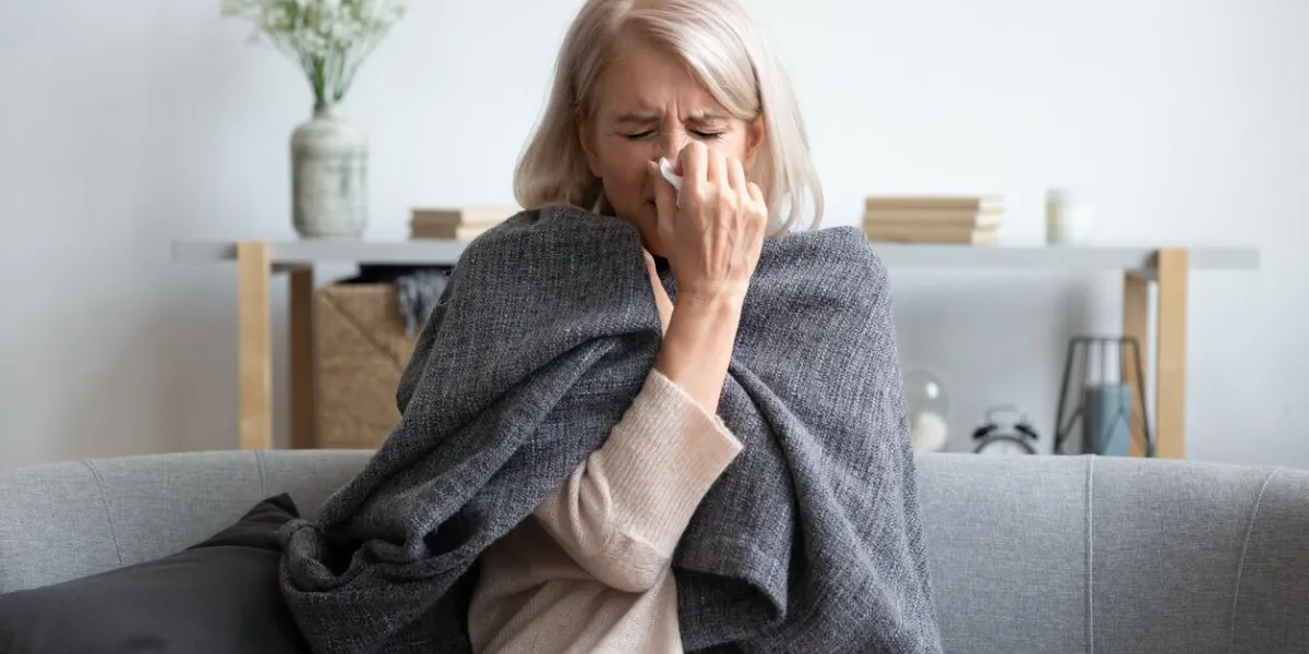 middle-aged 50s sick frozen woman seated on sofa in living room covered with warm plaid sneezing holding paper napkin blow out runny nose feels unhealthy, seasonal cold, weakened immune system concept