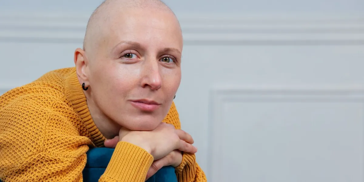 a bald woman cancer survivor with serene expression sitting in a chair, her hand resting on its arm, a white wall in the background
