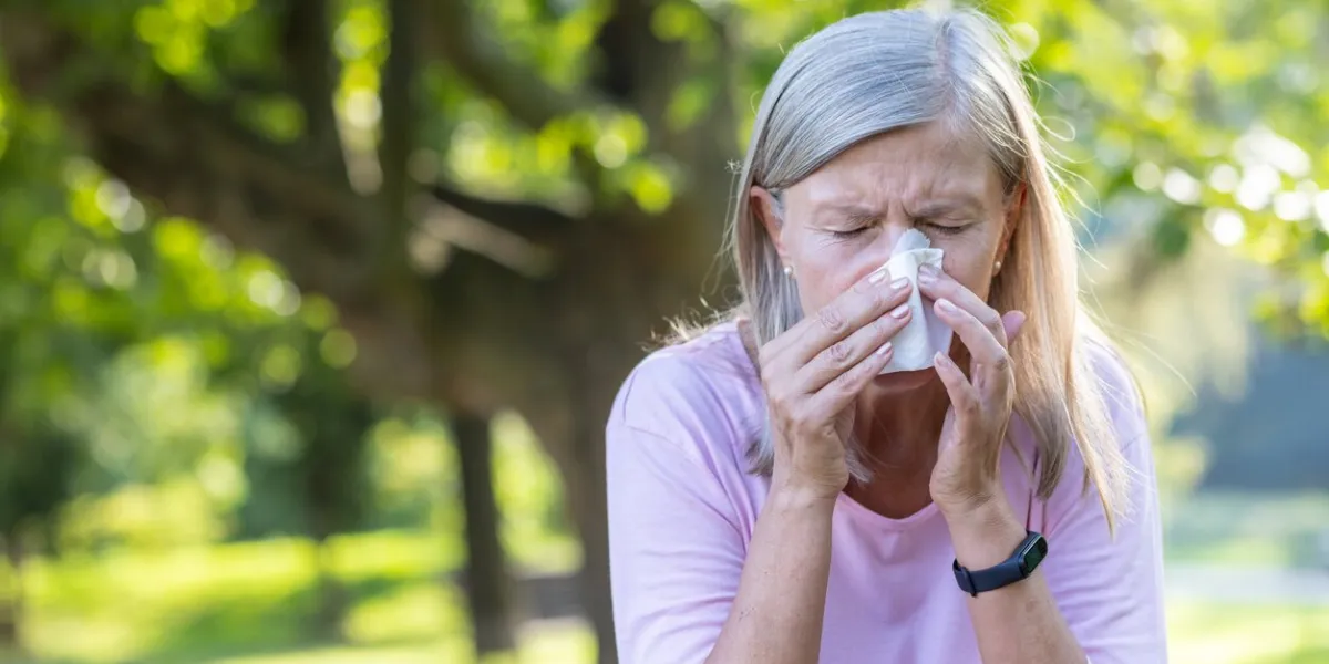close-up photo of a senior gray-haired woman standing outside in a park and wiping her nose with a napkin, suffering from a runny nose and seasonal allergies