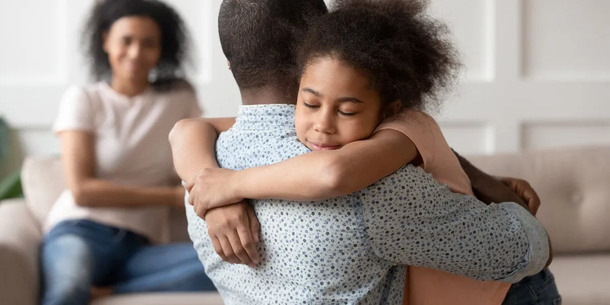 young african american man holding, embracing, comforting smiling happy calm black cute kid daughter, blurred mother sitting on couch on background, loving supporting family concept