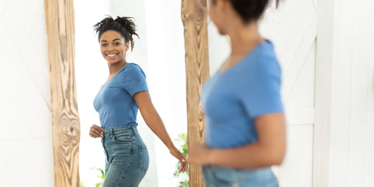 successful weight loss joyful african american girl after slimming smiling to her reflection in mirror standing at home staying fit, dieting and weight-loss concept selective focus