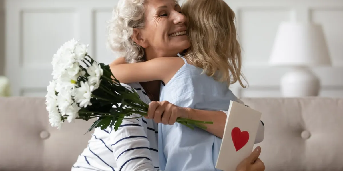 sincere old grandma hugs small attentive granddaughter express her gratitude for received surprise at birthday or 8-march woman day grandchild makes surprise for granny gave her flowers and postcard