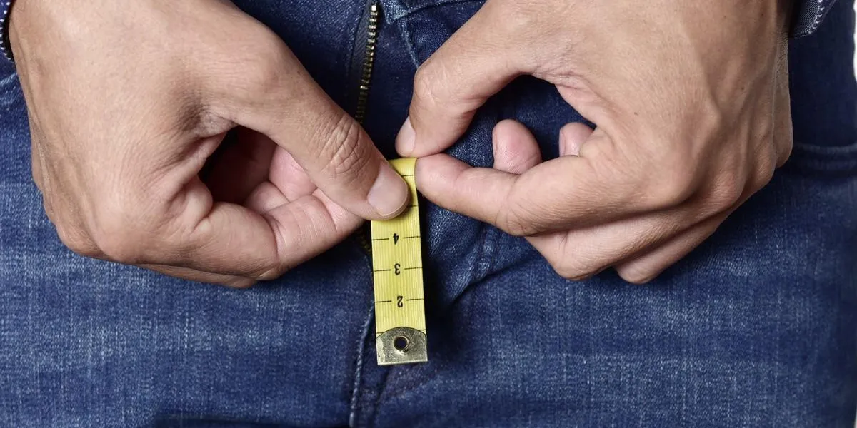 closeup of a young man holding a short piece of measuring tape that is popping up from the fly of his jeans