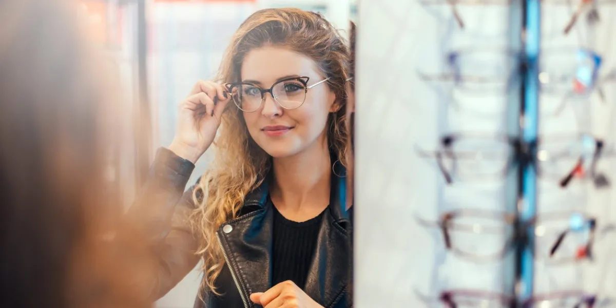 smiling young woman trying on glasses on mirror in optician