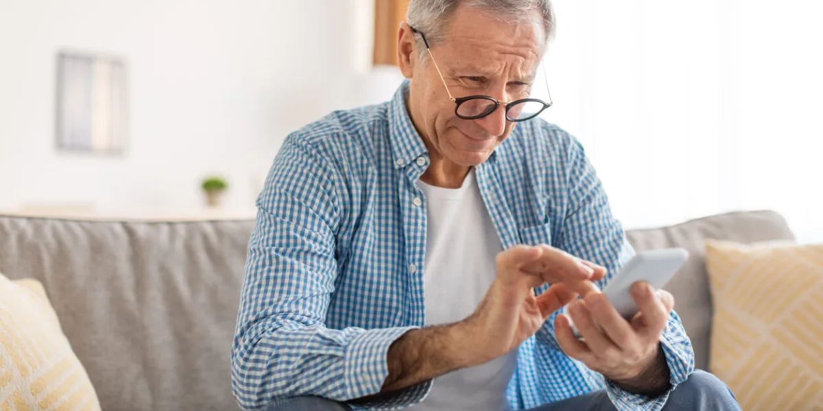 poor eyesight in older age, macular degeneration concept confused mature male in eyeglasses squinting eyes reading message on cell phone having ophtalmic issue problems with vision sitting on sofa