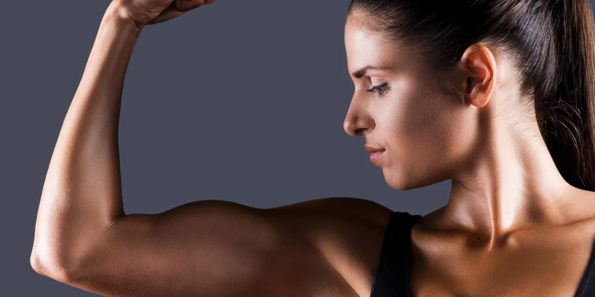 belle jeune femme sportive examinant son biceps en se tenant debout sur fond gris