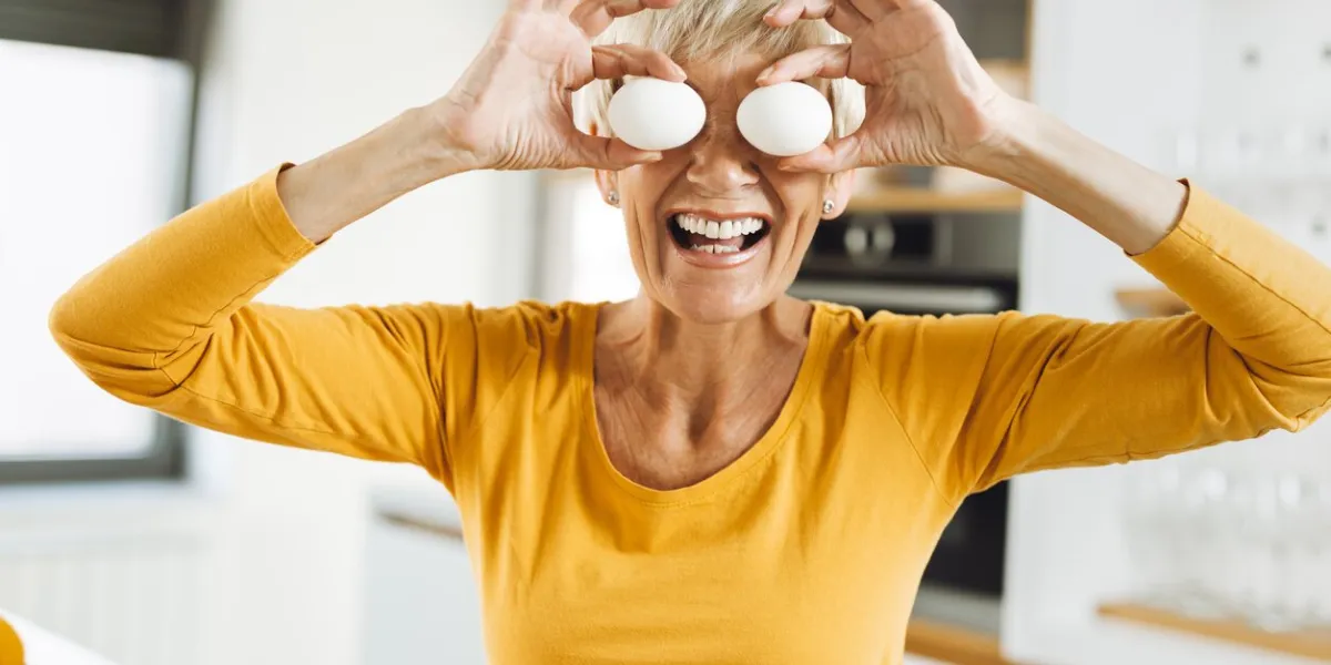happy senior woman covering her eyes with eggs and having fun in the kitchen