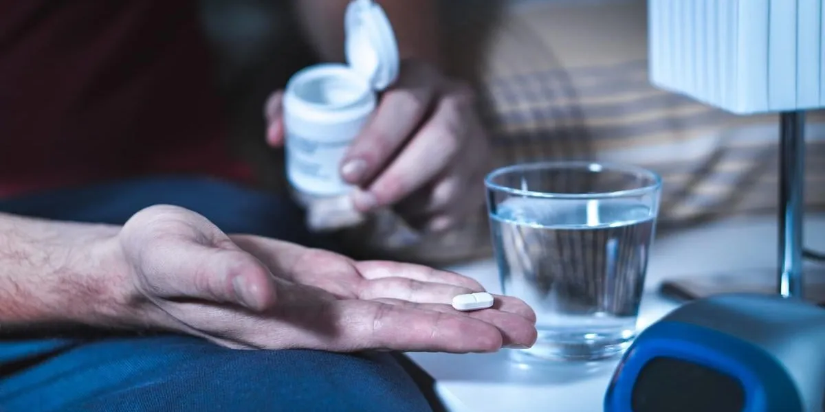 man sitting on bed about to take sleeping pill or night medicine suffering from insomnia holding tablet and bottle in hand glass of water on nightstand in bedroom