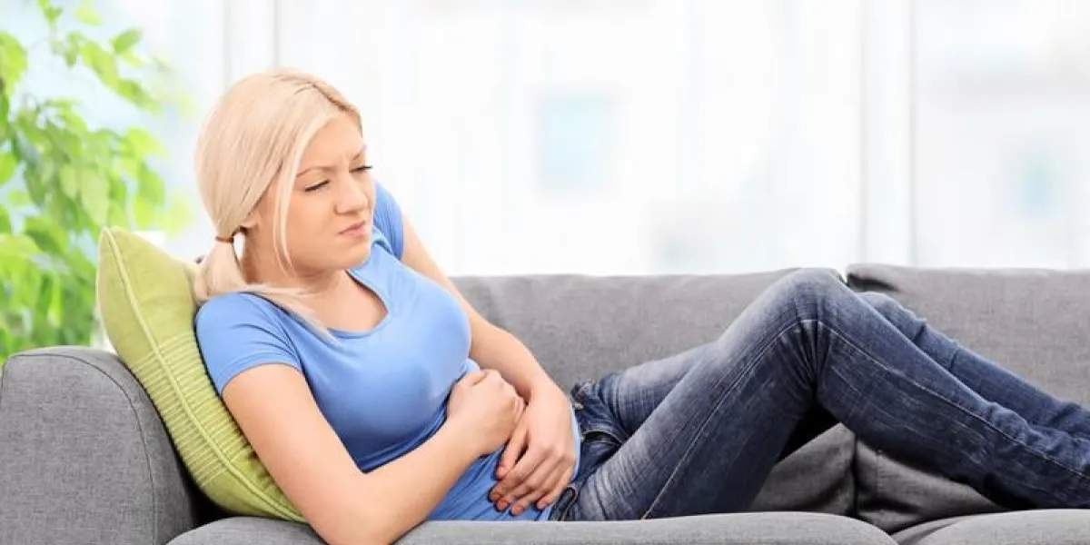 femme blonde avec une expression douloureuse assis sur un canapé gris à la maison avec ses mains placées sur son ventre