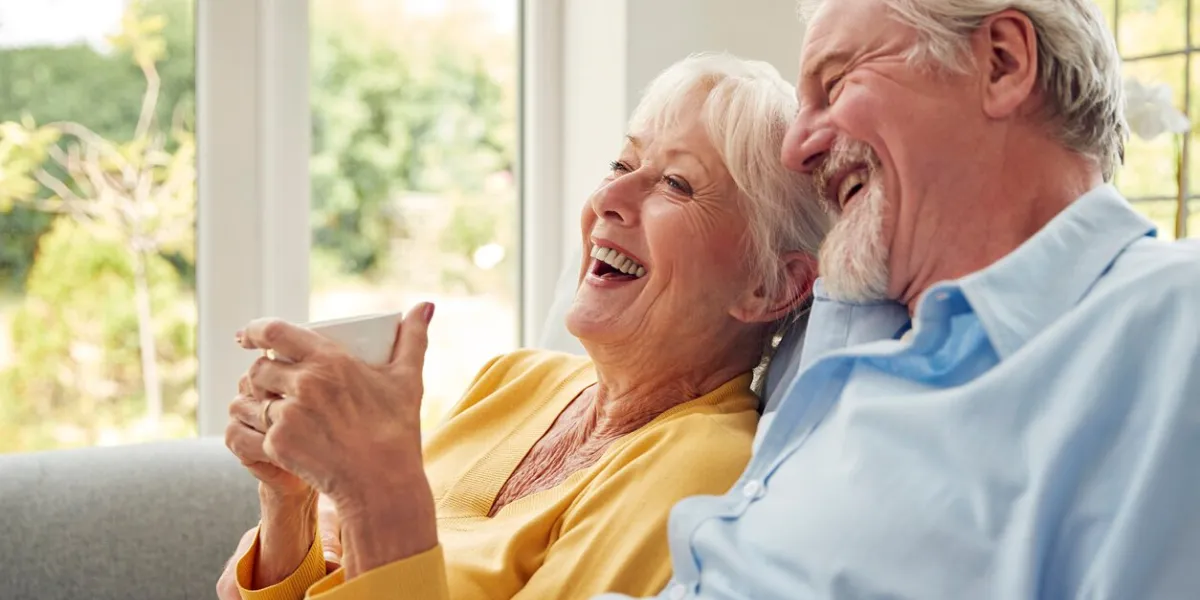 retired senior couple sitting on sofa at home drinking coffee and watching tv together