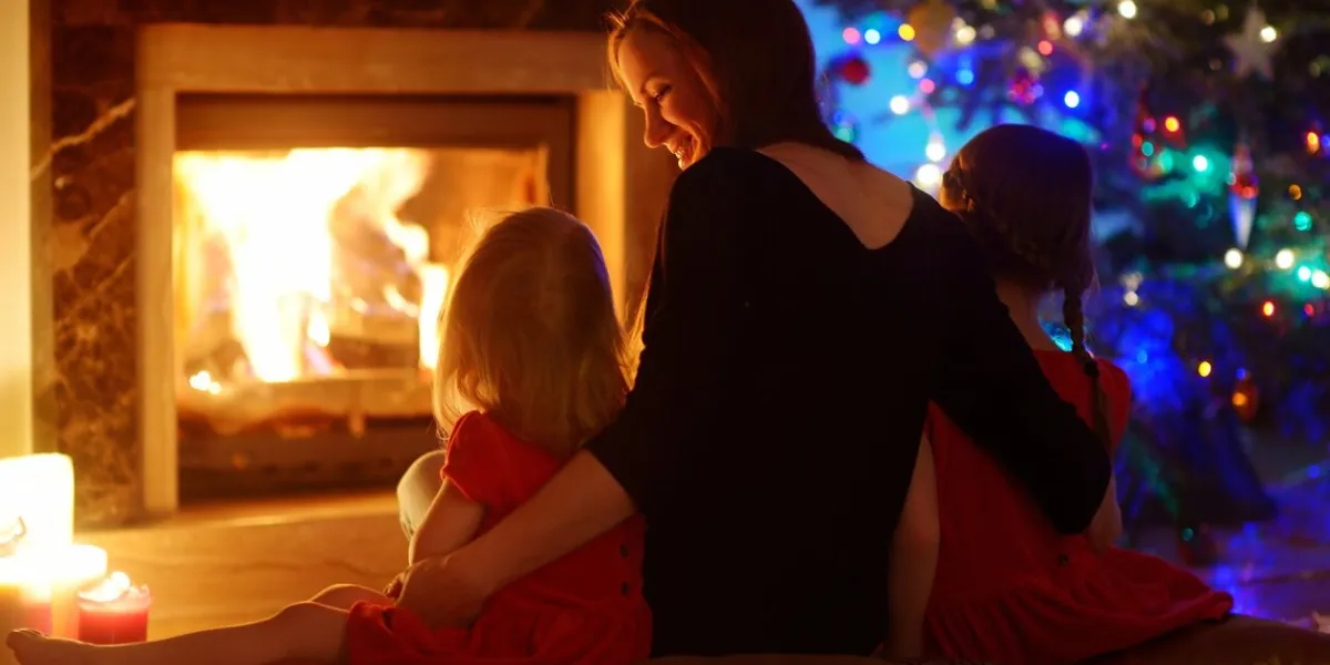 young mother and her two little daughters sitting by a fireplace in a cozy dark living room on christmas eve