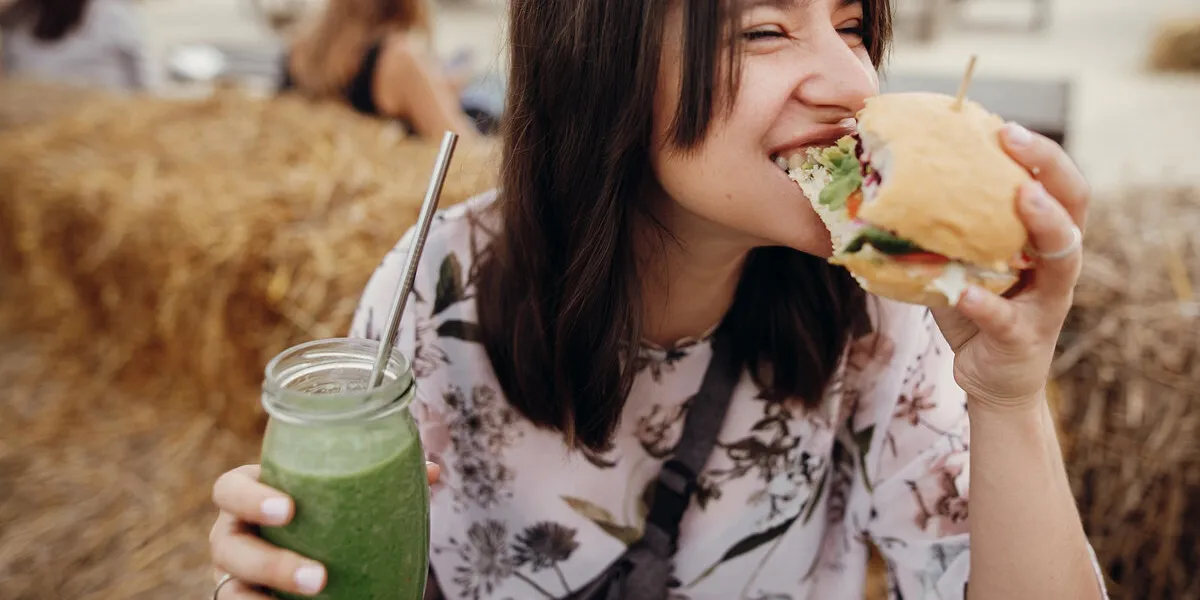 stylish hipster girl in sunglasses eating delicious vegan burger and holding smoothie in glass jar in hands at street food festival happy boho woman biting burger with drink in summer street
