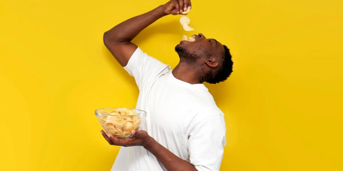 happy african american man holding plate of chips and eating snacks from potatoes on yellow isolated background, guy in white t-shirt eats fast food