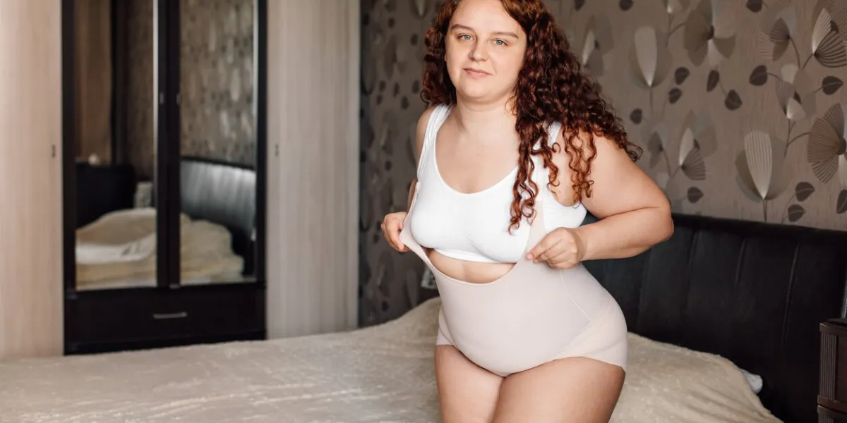 curly pleased overweight woman in white underwear stand near bed in bedroom and pull on slimming panties on sagging belly fat burning procedures of thick body weight loss process, diet and exercise