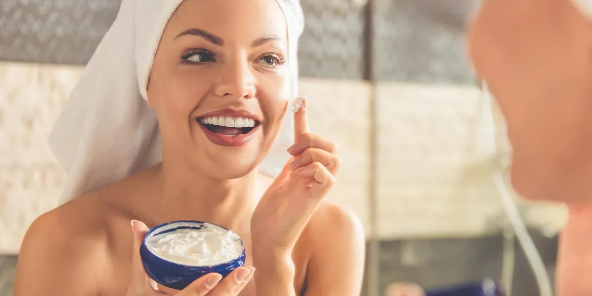 belle jeune femme en serviette de bain applique la crème sur son visage et souriant tout en regardant dans le miroir dans la salle de bain