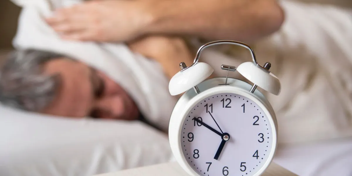sleeping man disturbed by alarm clock early morning sleepy young man covering ears with pillow as he looks at alarm clock in bed