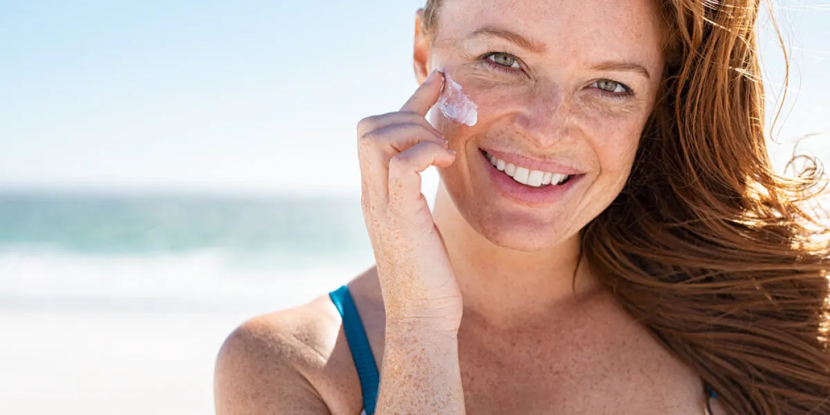 smiling young woman applying sunscreen lotion on face at beach, with copy space beautiful mature woman with red hair enjoying summer at sea portrait of happy girl using sunblock on her delicate skin with freckles and looking at camera