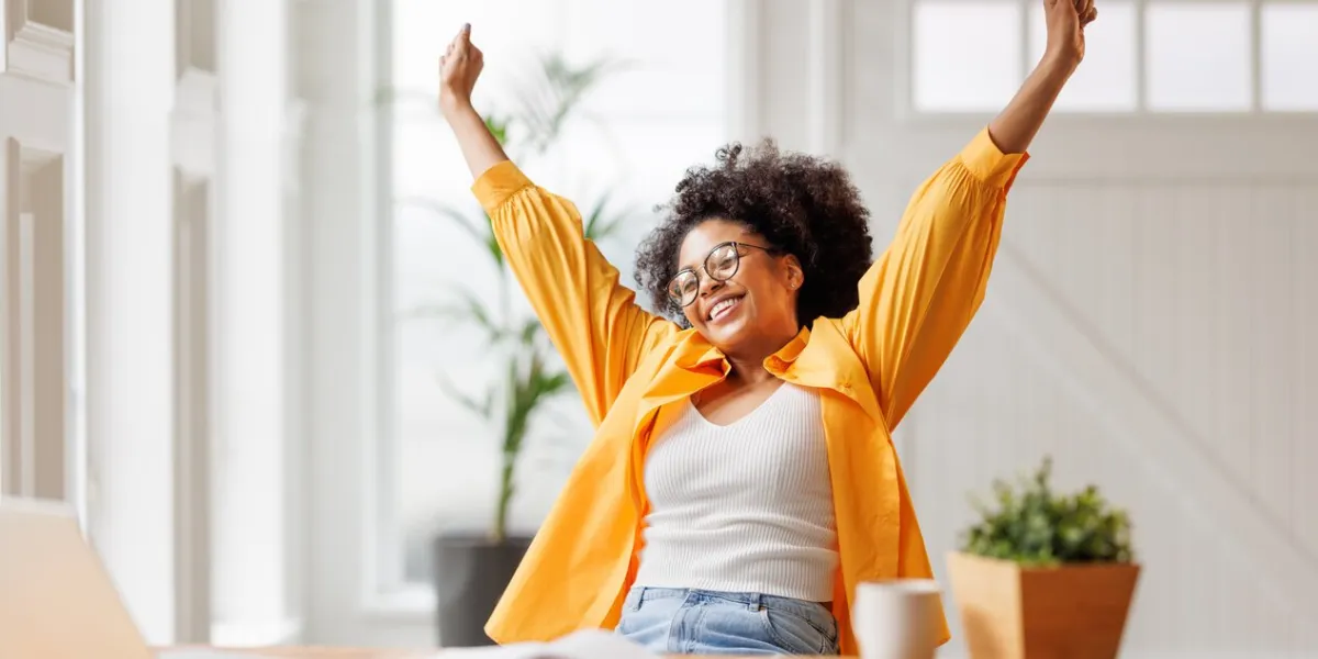joyful african american business woman freelancer  smiling and rejoices in victory while sitting at desk  and working at laptop screen after finishing project in home office