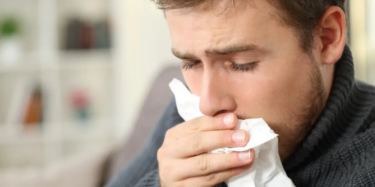 man coughing covering mouth with a tissue sitting on a couch in a house interior