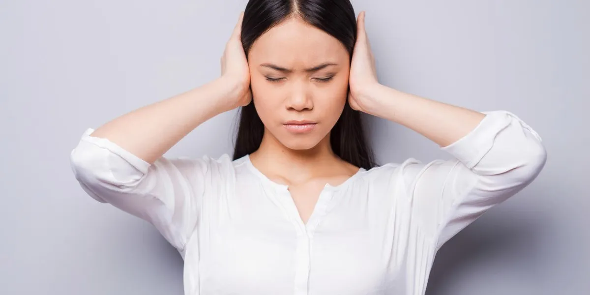beautiful young asian women covering ears with hands and keeping eyes closed while standing against grey background