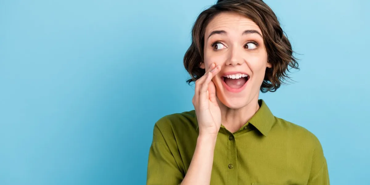 photo portrait of young pretty girl with short hair, telling secret information rumouring gossiping wearing green shirt isolated on blue color background