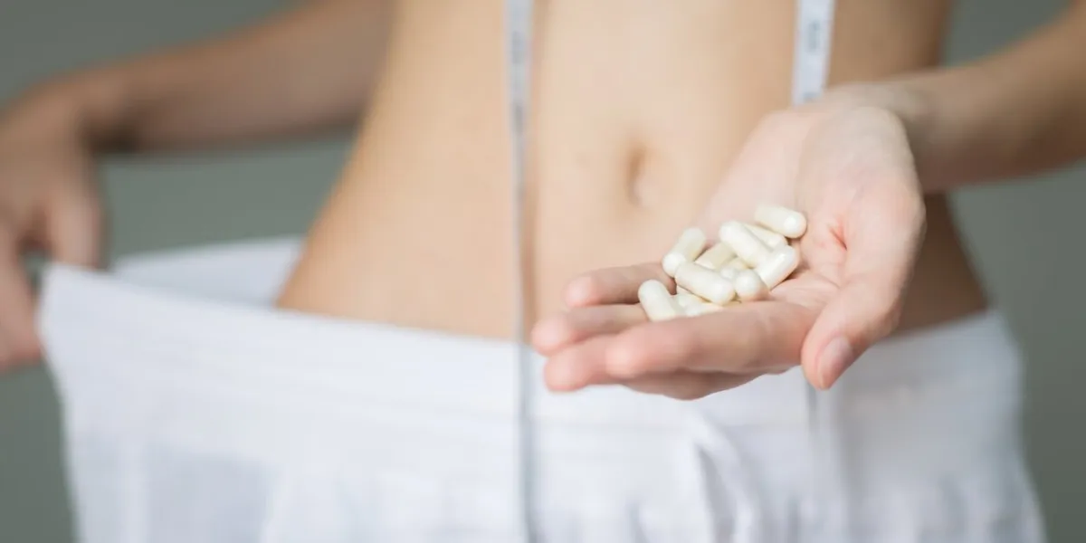 a woman showing the weight she loss with her oversize pants, meassuring tape, and diet fat burning suppliments