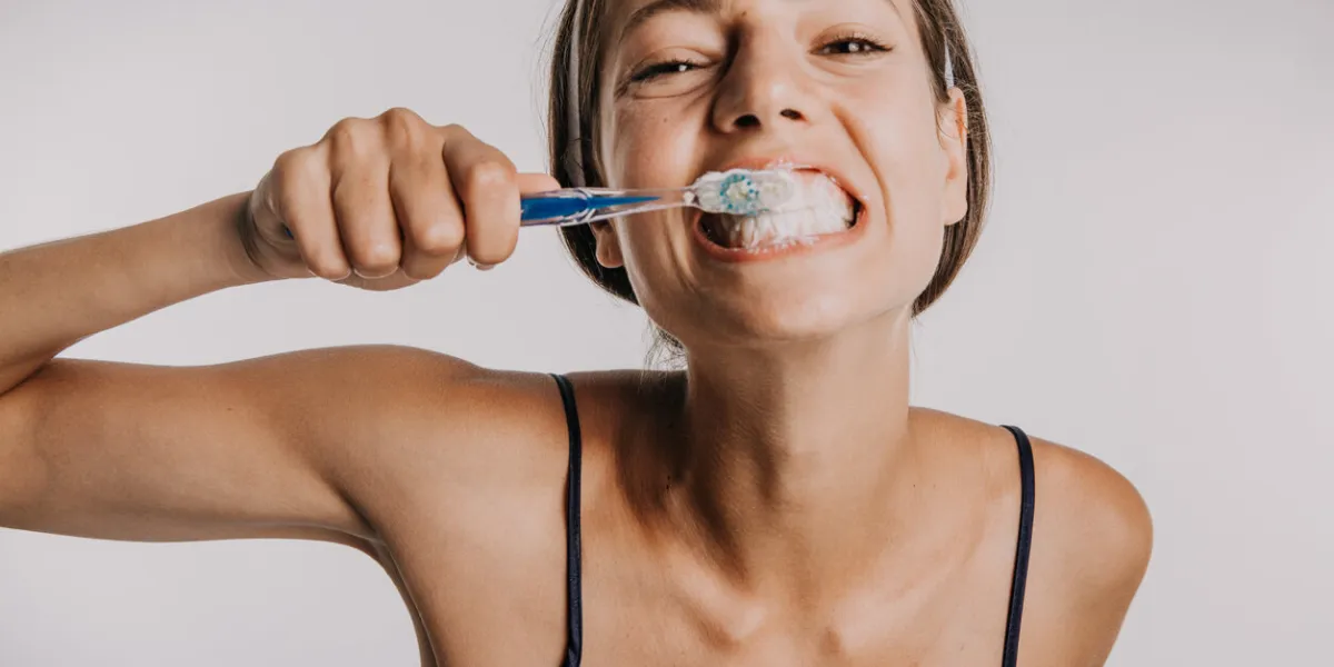 a funny photo of a cute young adult brushing her teeth wearing a nightgown, facing the camera isolated in a studio with a white background