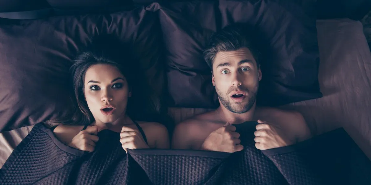 funny lovers man and woman lying in bed covered by sheets with wide opened eyes and mouth looking at camera, having shocked and scared expressions, afraid to be found in bed by someone who unexpectedly came home from work