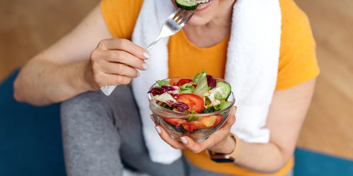 sports and nutrition concept fit senior woman sitting on yoga mat, eating fresh vegetable salad after domestic workout sporty aged lady enjoying her diet and exercising program