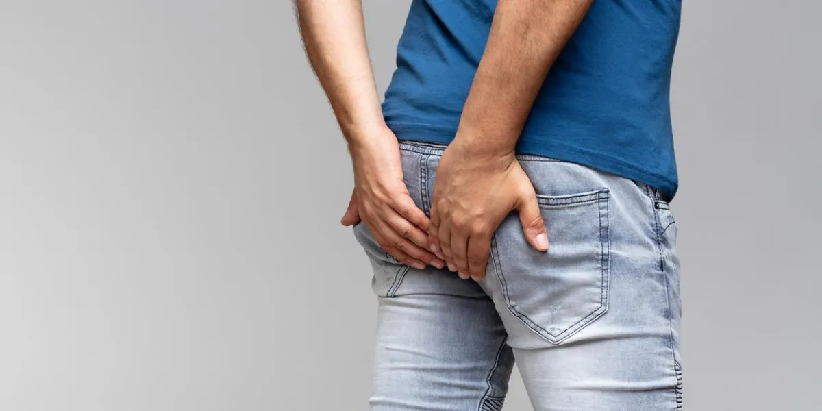 caucasian man in light blue t-shirt holding his buttocks in denim jeans on a gray background with copy space