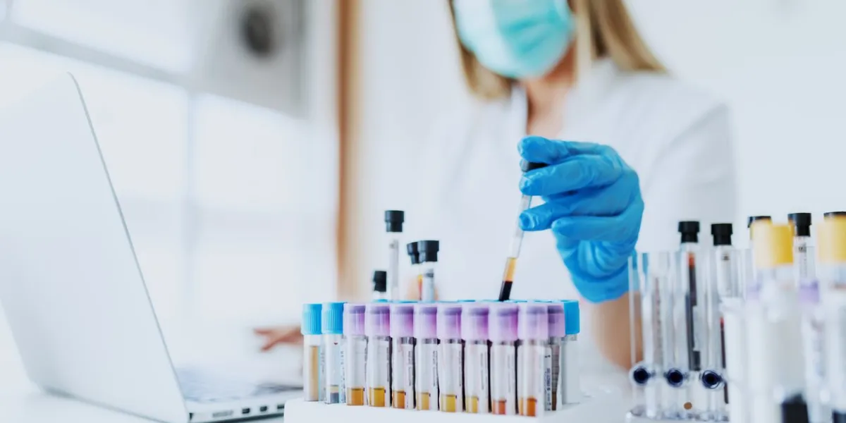 close up of lab assistant in uniform, with mask and rubber gloves holding test tube with blood sample while sitting on chair and typing on laptop selective focus on test tubes