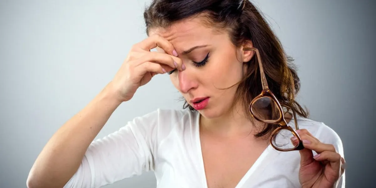 séduisante jeune femme avec un mal de tête tenant ses lunettes dans une main comme elle pince le pont de son nez avec une expression peinée, la tête et les épaules sur gris