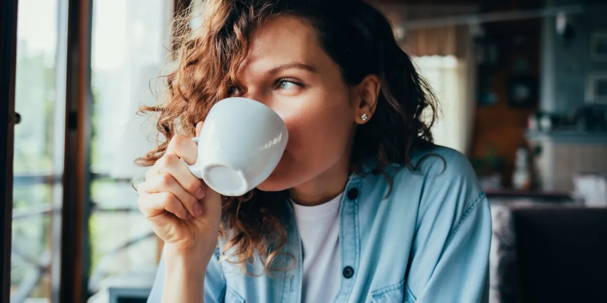 portrait of happy beautiful young woman wearing blue shirt drinking coffee sitting at table in restaurant looking out the window on summer day