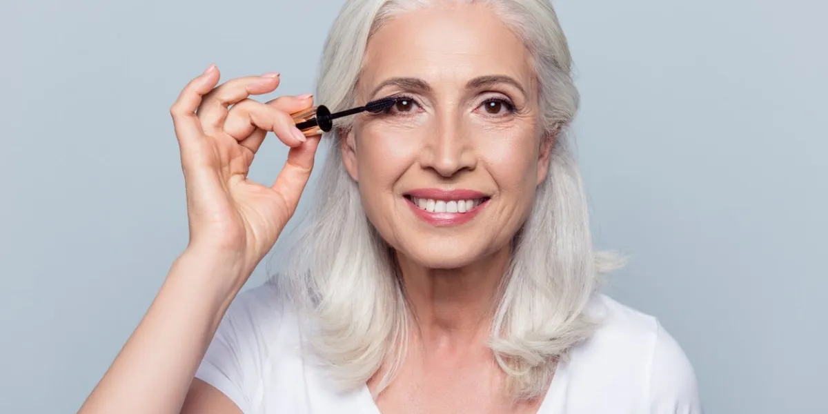 nice, delighted, caucasian, charming, pretty, aged woman applying black mascara, dye eyelashes using tassel, making, doing professional make up over grey background