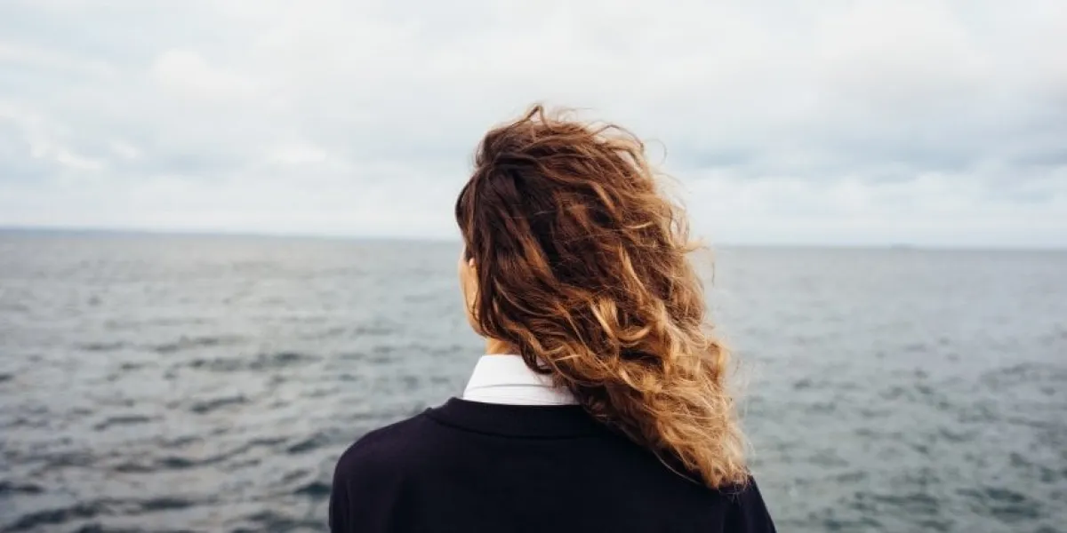 rear view of young woman looking at overcast sky and gray sea female with red curly hair standing alone thinking at the background of seascape