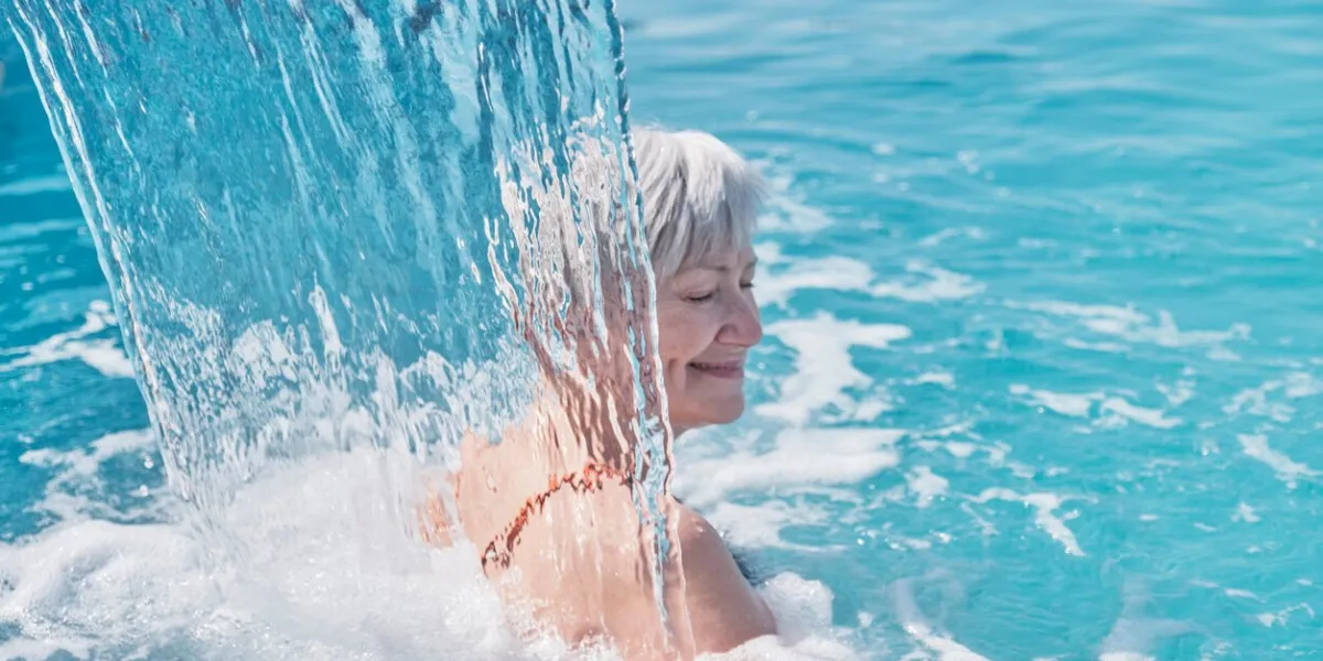 senior caucasian smiling woman with gray hair enjoying falling on her shoulders flow of water hydromassage in outdoor thermal swimming pool active elderly concept blurry movement