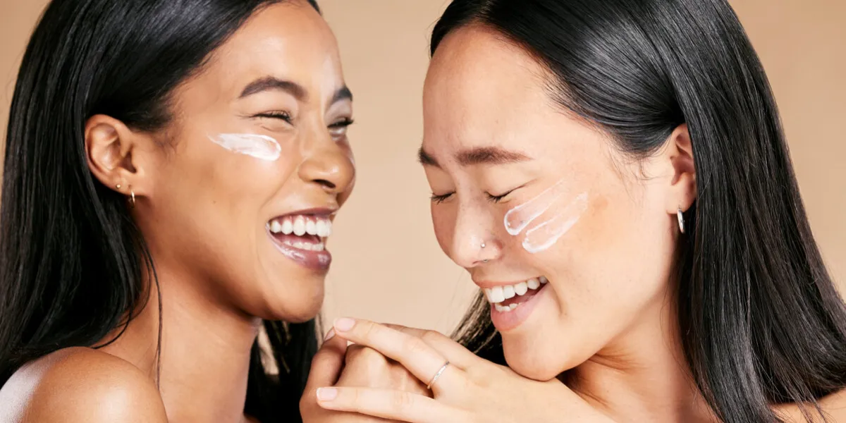 happy, beauty and skincare women with cream for face isolated on a beige studio background wellness, smile and asian model friends with sunscreen for love of cosmetics and dermatology on a backdrop