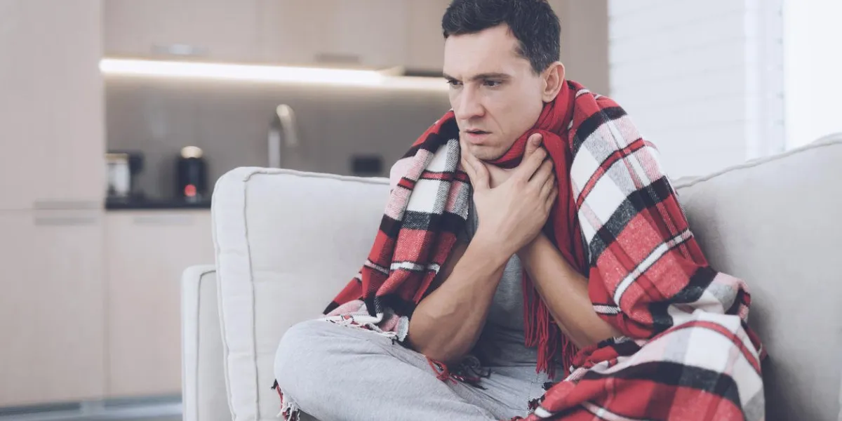 a man with a cold sits on the couch, hiding behind a red rug he's holding his throat because it hurts badly