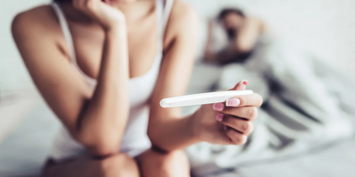 cropped image of young woman is sitting on the edge of bed with pregnancy test in hand man is sleeping on the background