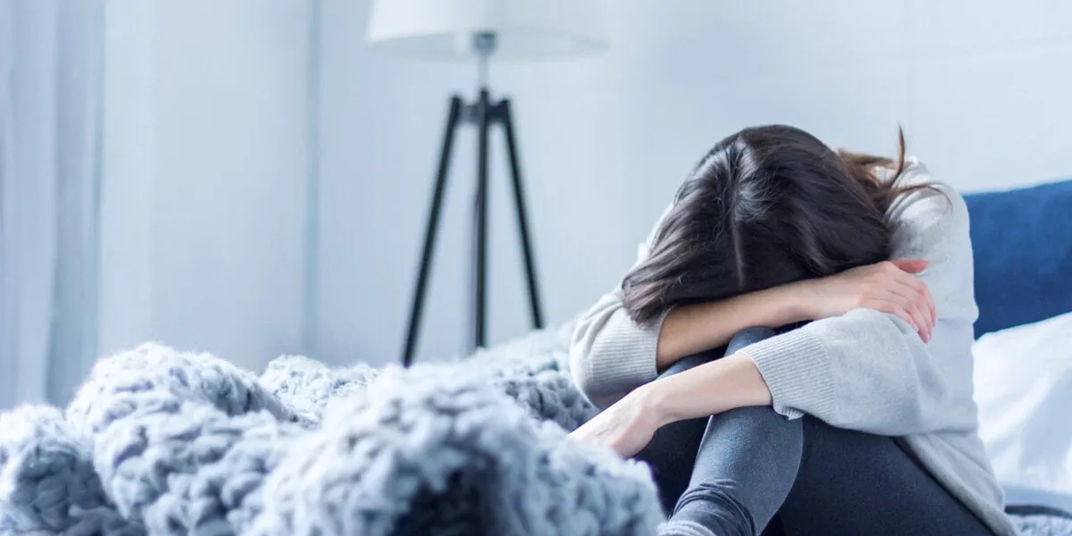 young woman in gray clothes is sitting curled up on bed at home upset brunette girl is suffering, crying depression because of loneliness, stress, problems psychological disorders concept