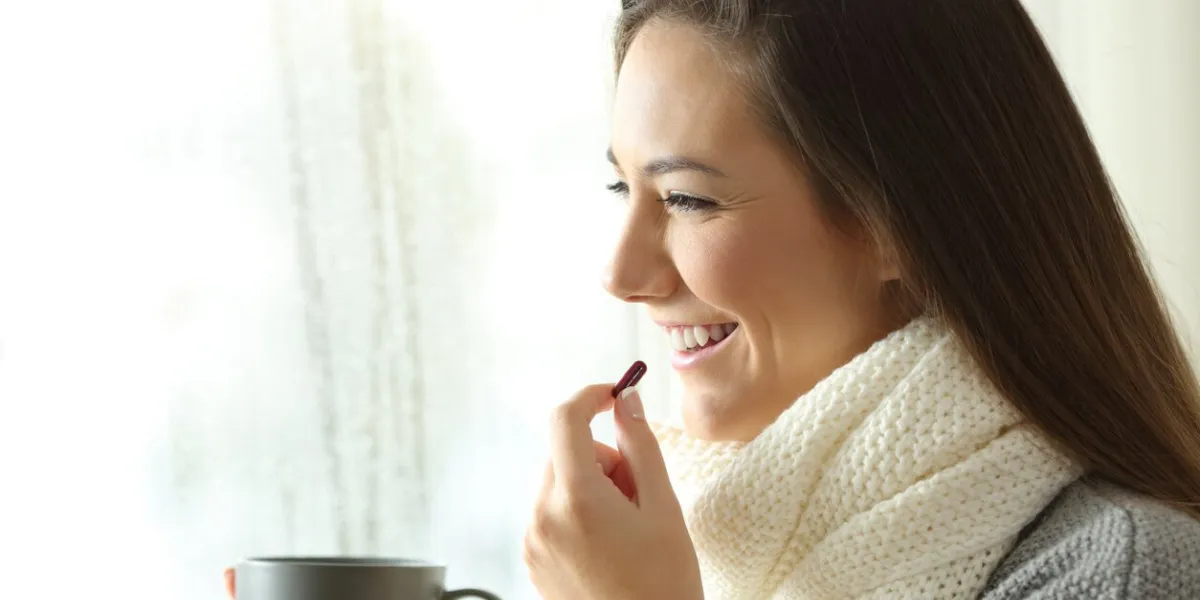 side view portrait of a happy woman taking a pill and looking through a window in a rainy day of winter