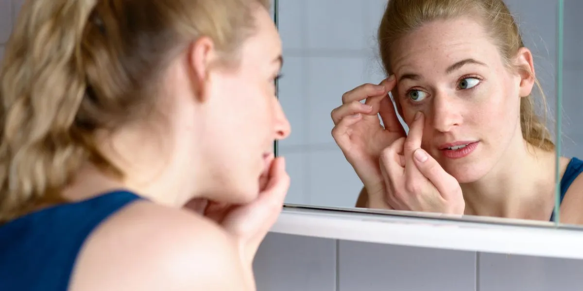 over the shoulder view of a beautiful young woman worried about aging while looking in the mirror at the skin around her eyes
