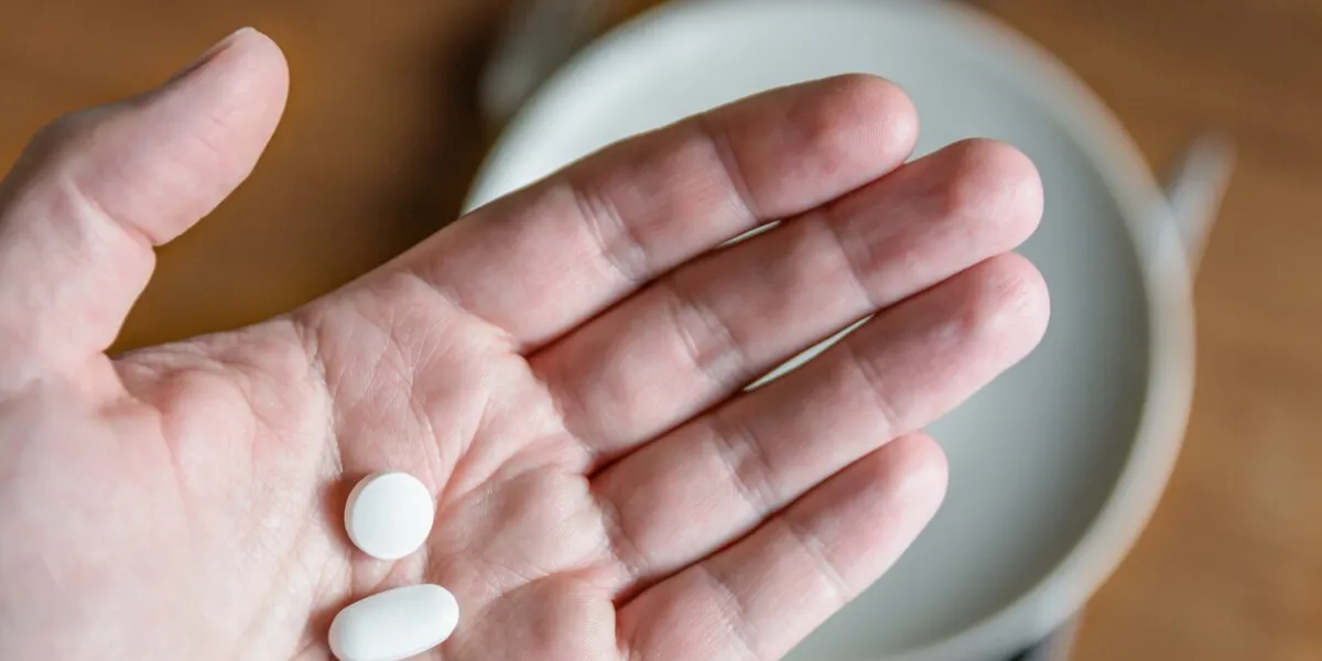 close-up view of a white male hand holding two white pills in the palm above a blurry background showing a dining table with plate, cutlery and glass