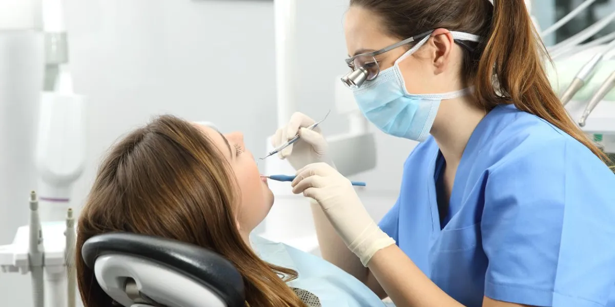 dentist wearing eyeglasses gloves and mask examining a patient teeth with a dental probe and a mirror in a clinic box with equipment in the background