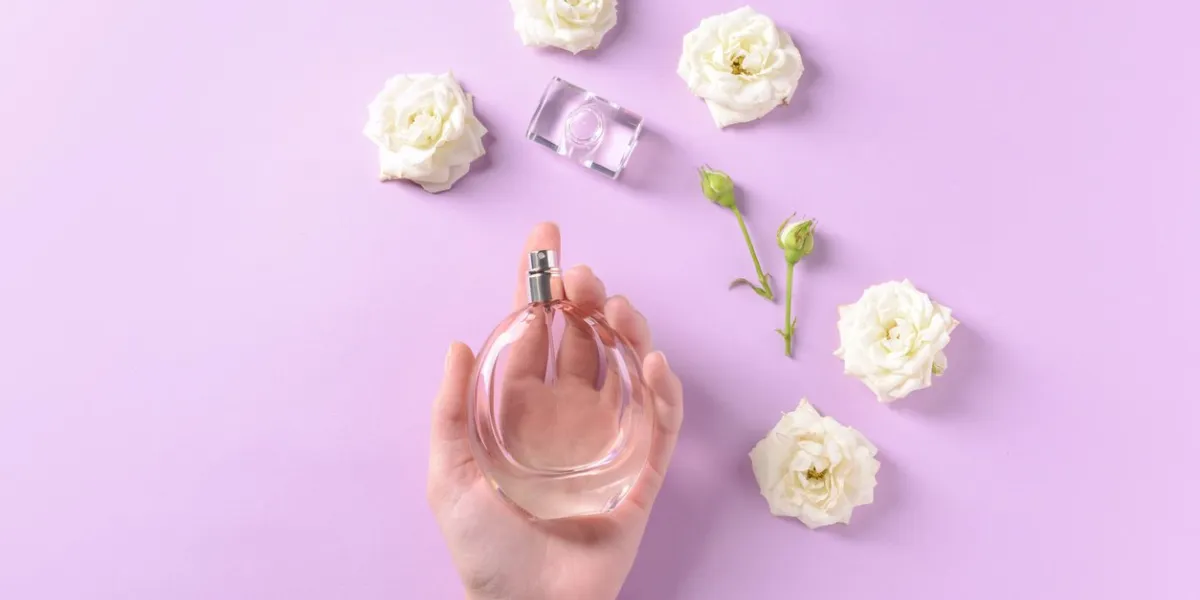 female hand with bottle of perfume and flowers on color background