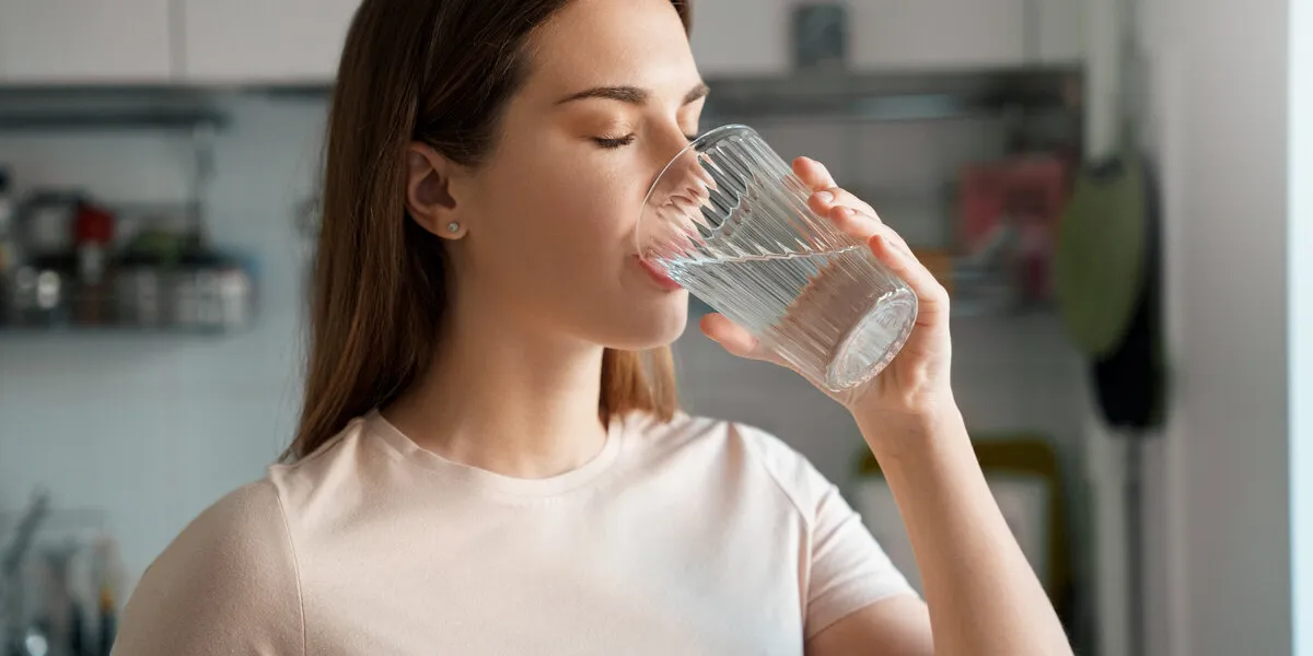 thirsty young woman drinking fresh water from glass home office kitchen interior headshot portrait dehydration prevention, normal bowel function and balance of body maintenance concept